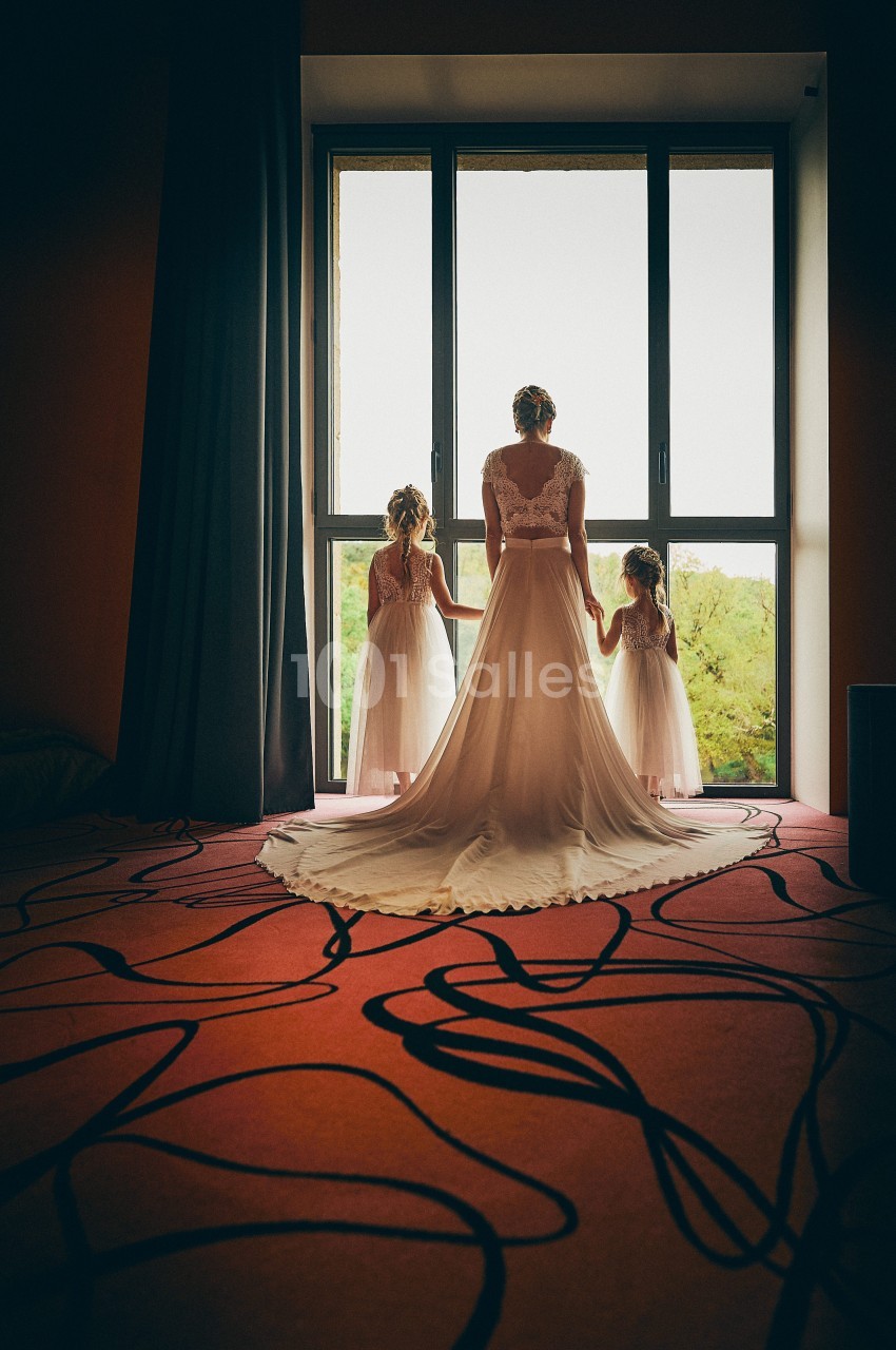 Une mariée en robe blanche tient la main de deux enfants devant une grande fenêtre donnant sur un paysage verdoyant.