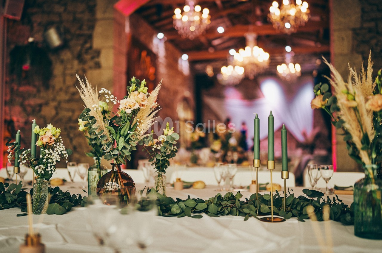 Table décorée avec des bouquets de fleurs, chandeliers verts et guirlande végétale dans une salle éclairée par des lustres.