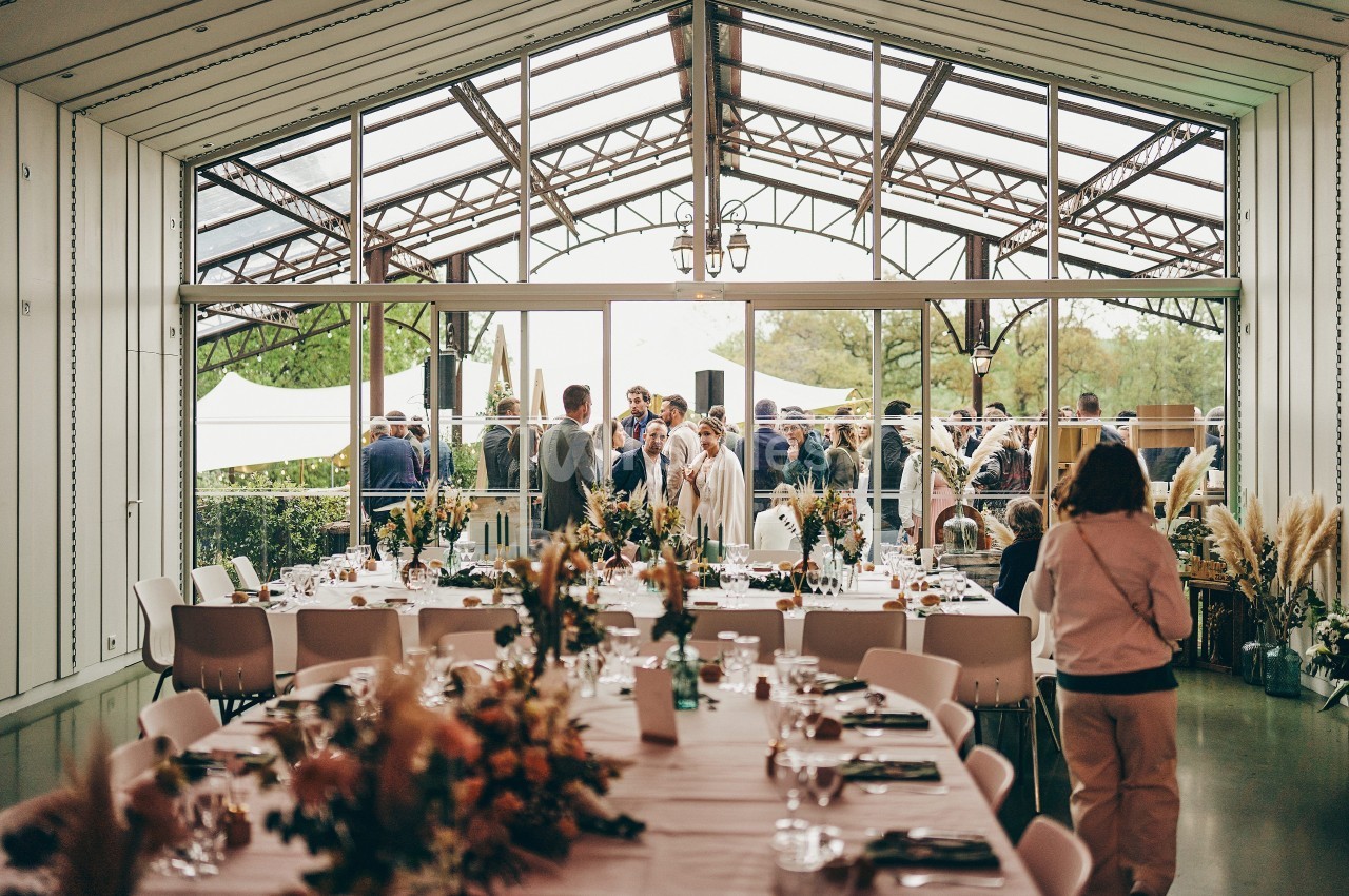 Salle de réception lumineuse avec tables décorées, vue sur une terrasse où des invités sont rassemblés.