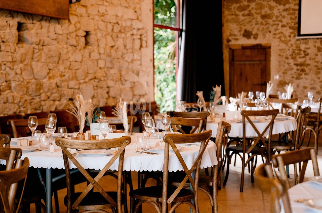 Salle de réception avec des tables rondes dressées, nappes blanches et chaises en bois, murs en pierre apparente.