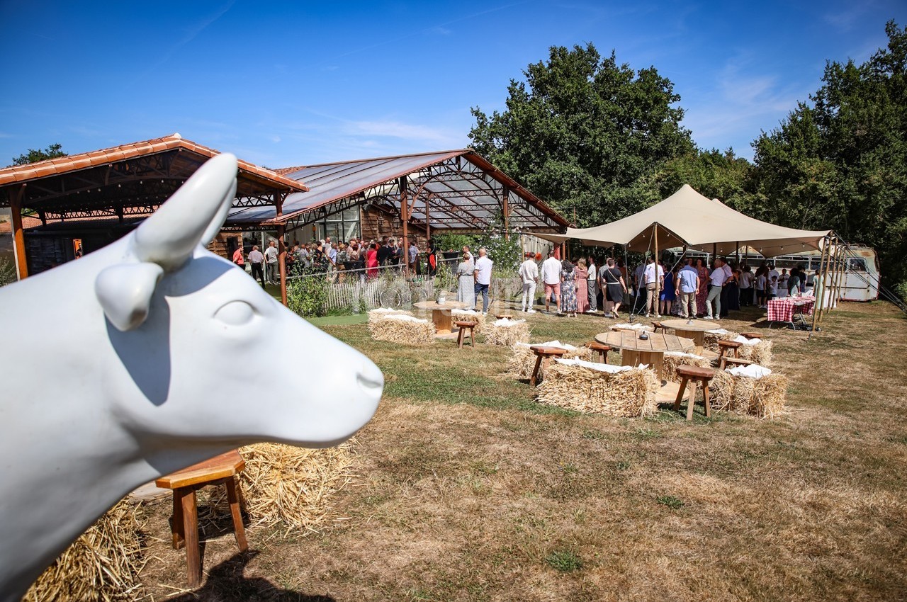 Statue de vache blanche au premier plan, avec des stands et des visiteurs dans un espace extérieur en arrière-plan.