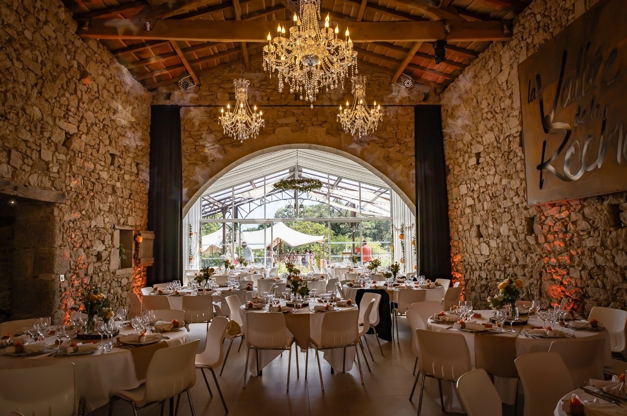 Salle de réception en pierre avec tables décorées, lustres suspendus et grande baie vitrée donnant sur l'extérieur.