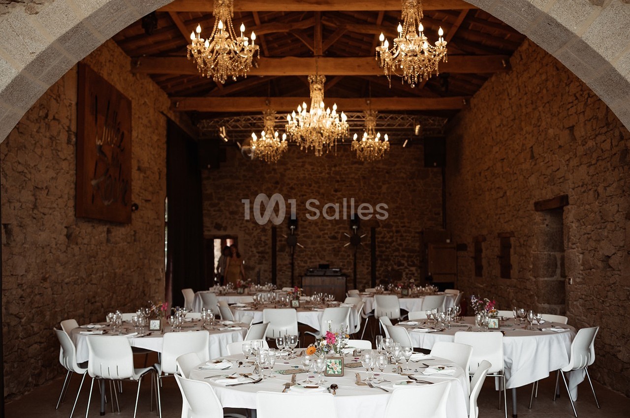 Salle en pierre avec tables rondes dressées pour un événement, éclairée par des lustres suspendus au plafond en bois.