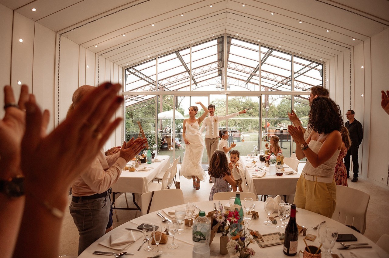 Un couple en tenue de mariage entre dans une salle lumineuse sous les applaudissements des invités.