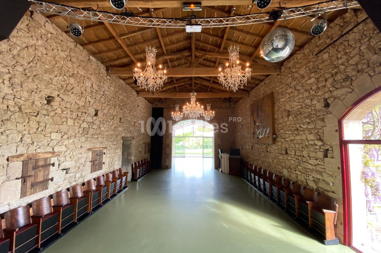 Salle en pierre avec plafond en bois, lustres suspendus, rangées de chaises et grande baie vitrée donnant sur l'extérieur.