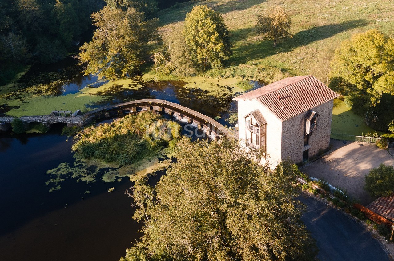 Vue aérienne d'un moulin en pierre près d'une rivière avec un pont en bois entouré de végétation.