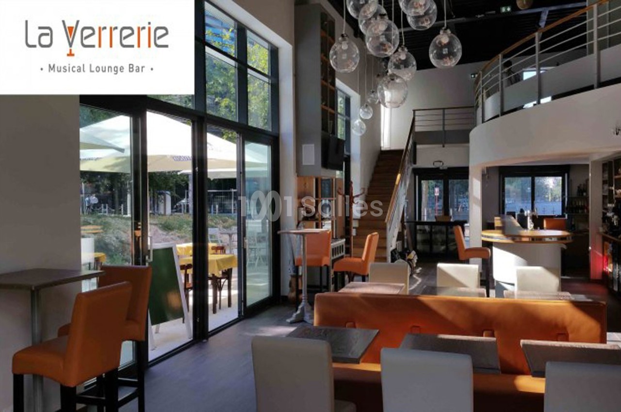 Intérieur lumineux d'un bar lounge avec canapés orange, tables, baies vitrées et mezzanine en arrière-plan.