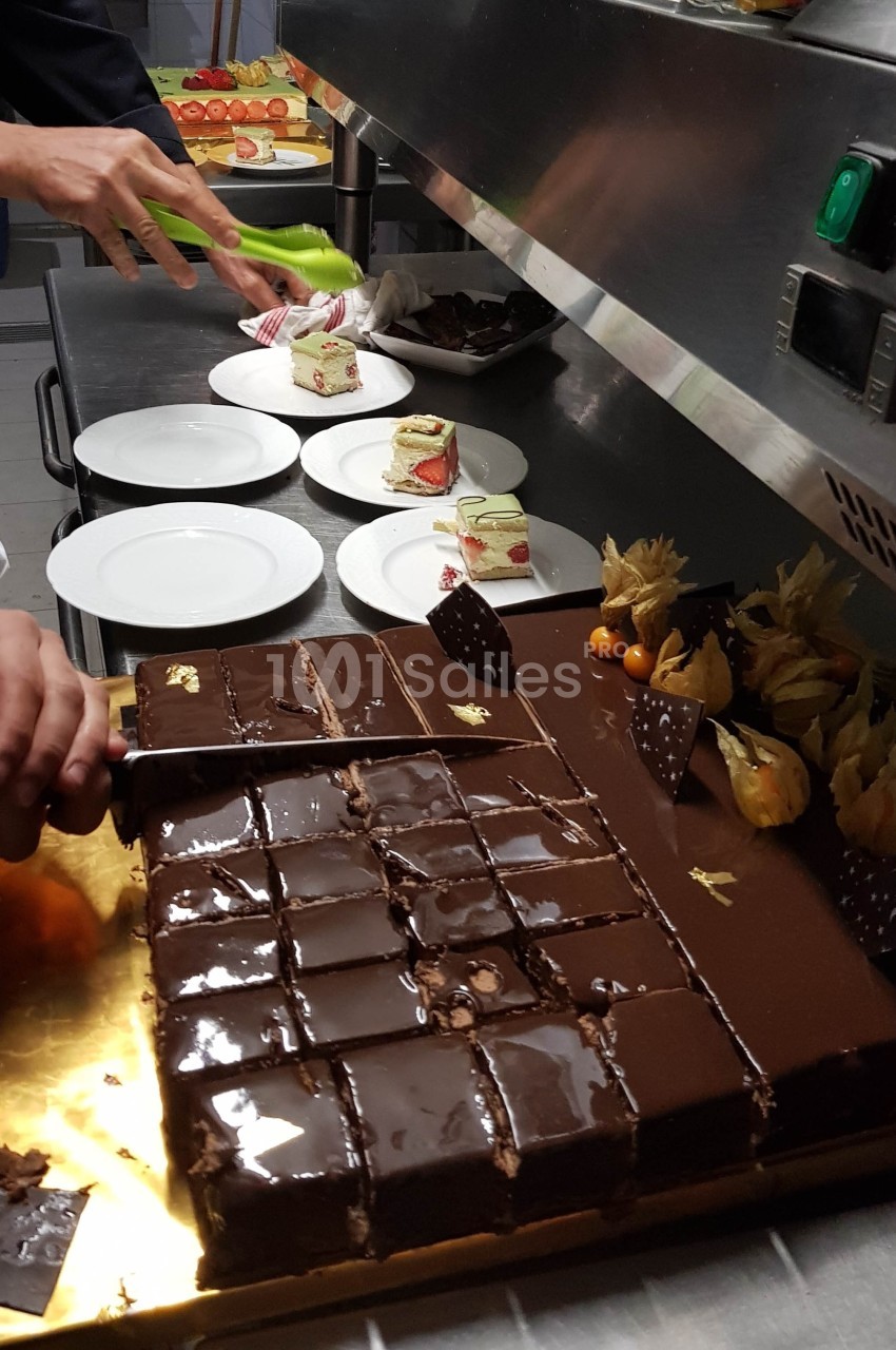 Découpe d'un gâteau au chocolat brillant en cuisine, avec des desserts et assiettes préparés en arrière-plan.