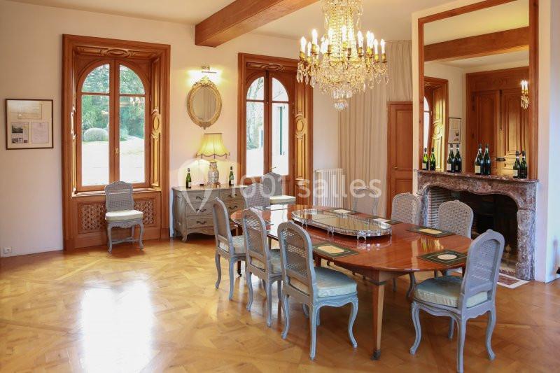 Salle à manger élégante avec table en bois, chaises classiques, lustre, cheminée et grandes fenêtres donnant sur un jardin.