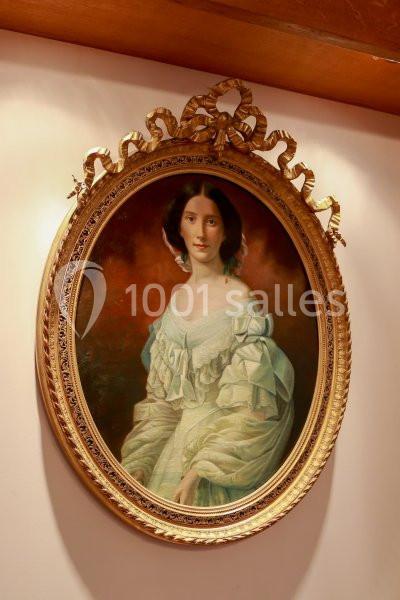Portrait d'une femme en robe blanche dans un cadre ovale doré, accroché sur un mur beige.
