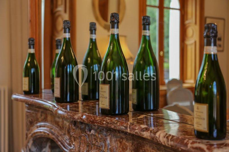 Bouteilles de champagne alignées sur une cheminée en marbre dans une pièce lumineuse avec des fenêtres en arrière-plan.