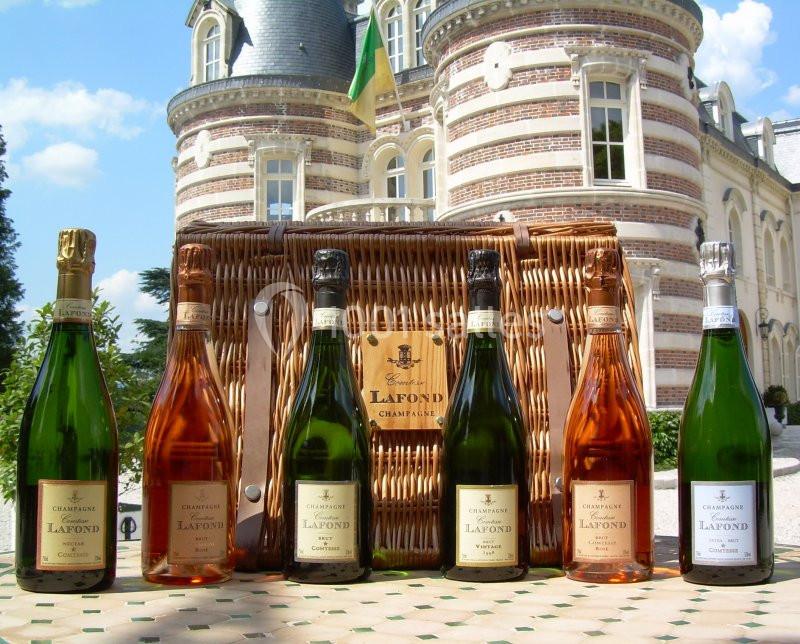 Six bouteilles de champagne devant un panier en osier et un bâtiment en briques avec des détails architecturaux élégants.