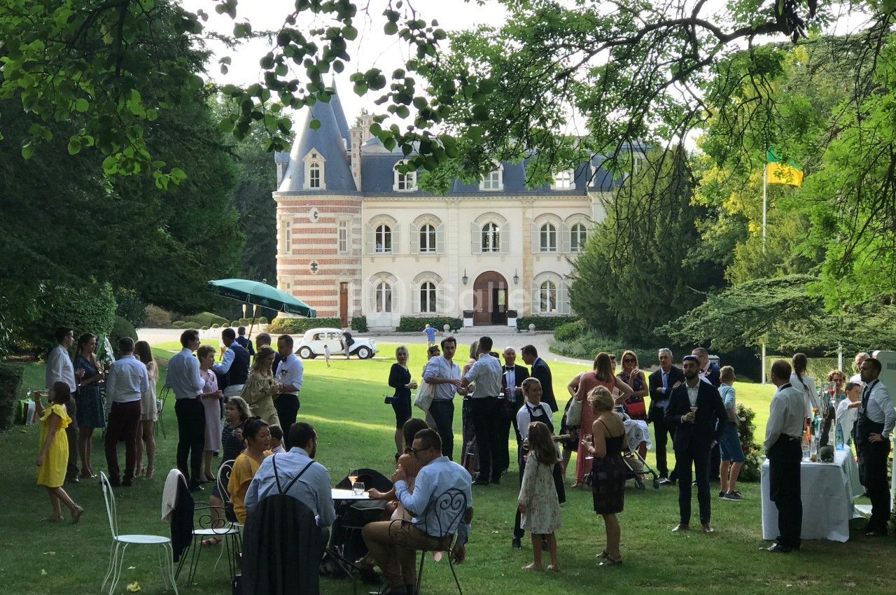 Groupe de personnes rassemblées dans un jardin devant un bâtiment de style château, lors d'un événement en plein air.
