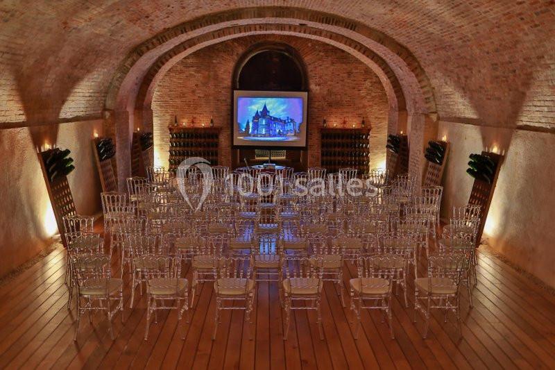 Salle voûtée en briques avec des chaises transparentes alignées face à un écran projetant une image de château.