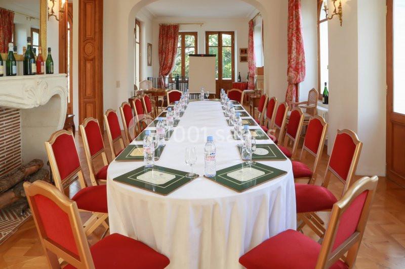 Salle de réunion lumineuse avec une grande table ovale dressée, entourée de chaises rouges et décorée de rideaux fleuris.