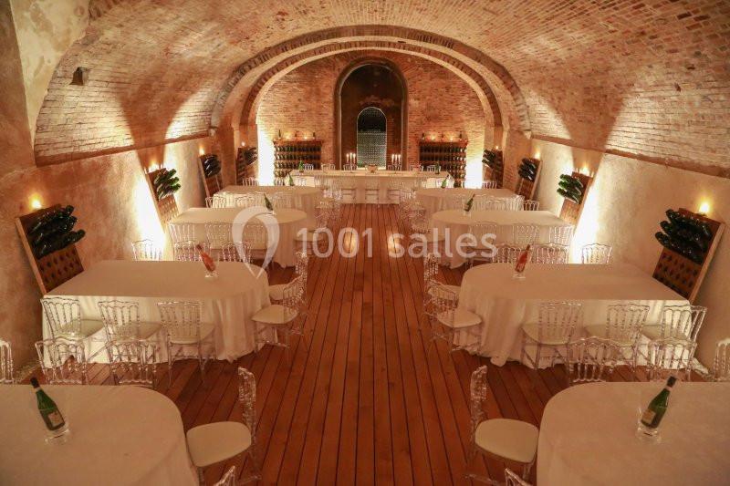Salle voûtée en briques avec tables rondes dressées, chaises transparentes et bouteilles de vin exposées sur les murs.