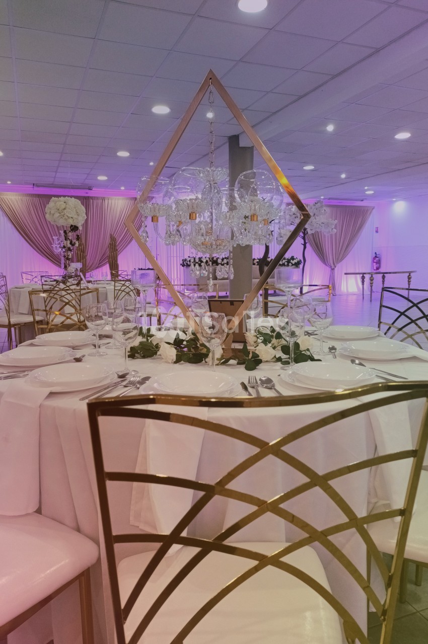 Salle de réception décorée avec des tables élégantes, chaises dorées, nappes blanches et éclairage rose tamisé.
