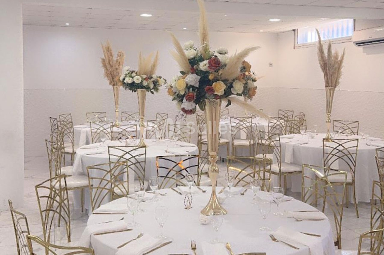 Salle de réception avec tables rondes dressées, décorées de centres de table floraux et chaises dorées.