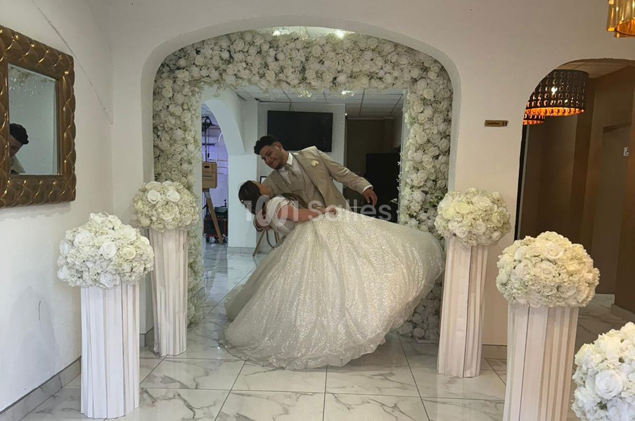 Un couple en tenue de mariage danse sous une arche décorée de fleurs blanches dans une salle lumineuse.