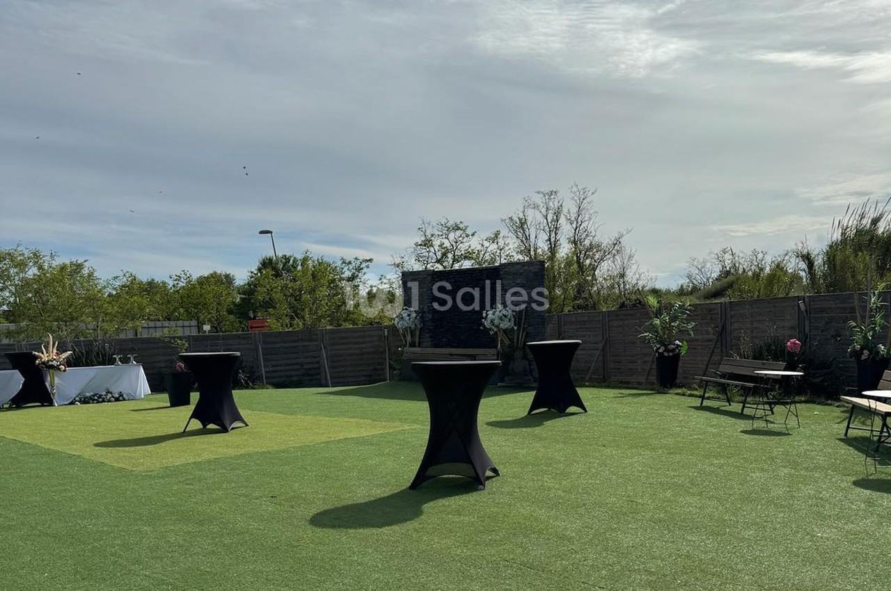 Espace extérieur avec pelouse synthétique, tables hautes noires et quelques plantes en pot, sous un ciel partiellement…