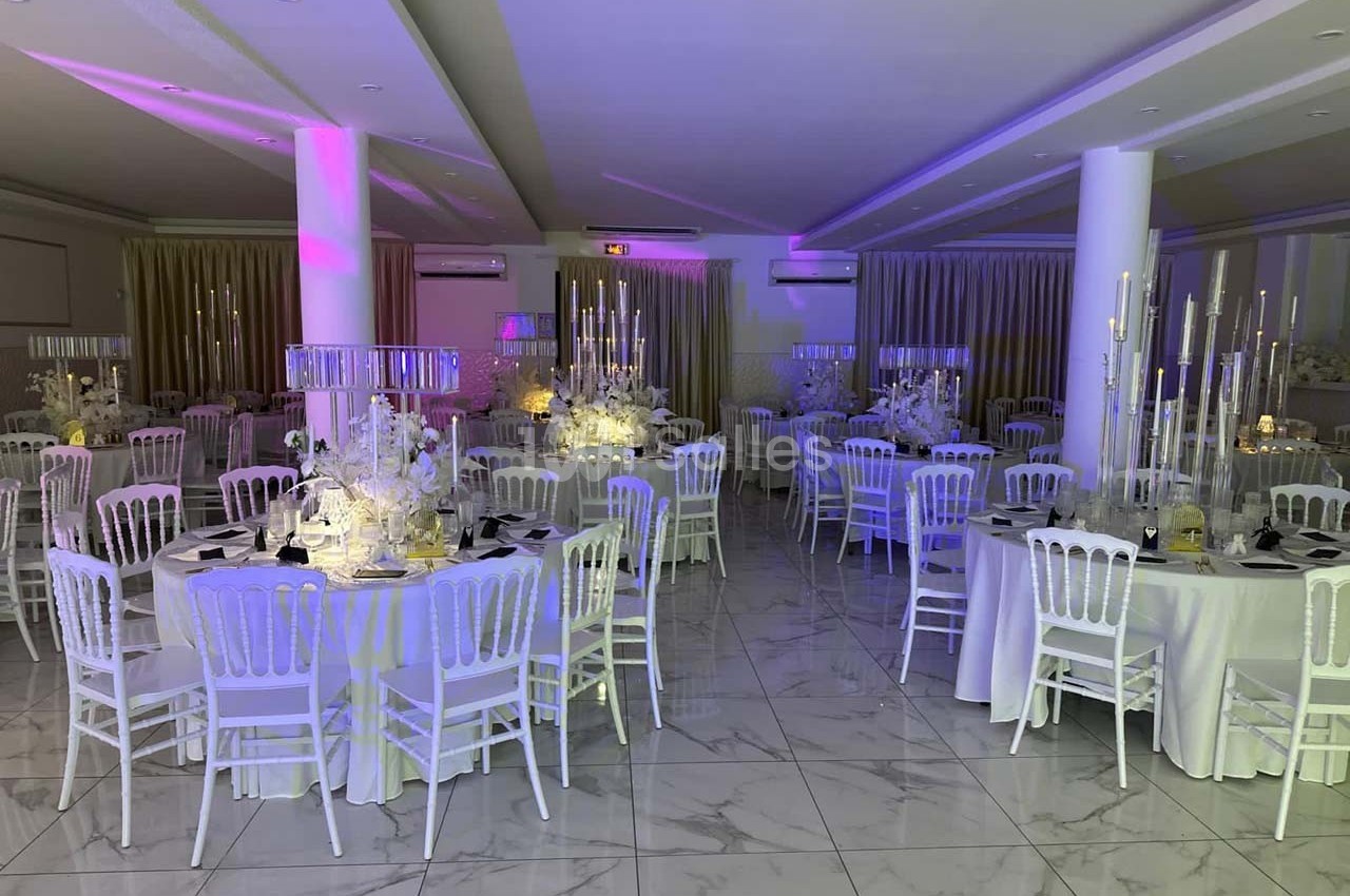 Salle de réception élégamment décorée avec des tables rondes, des chaises blanches et des chandeliers illuminés.
