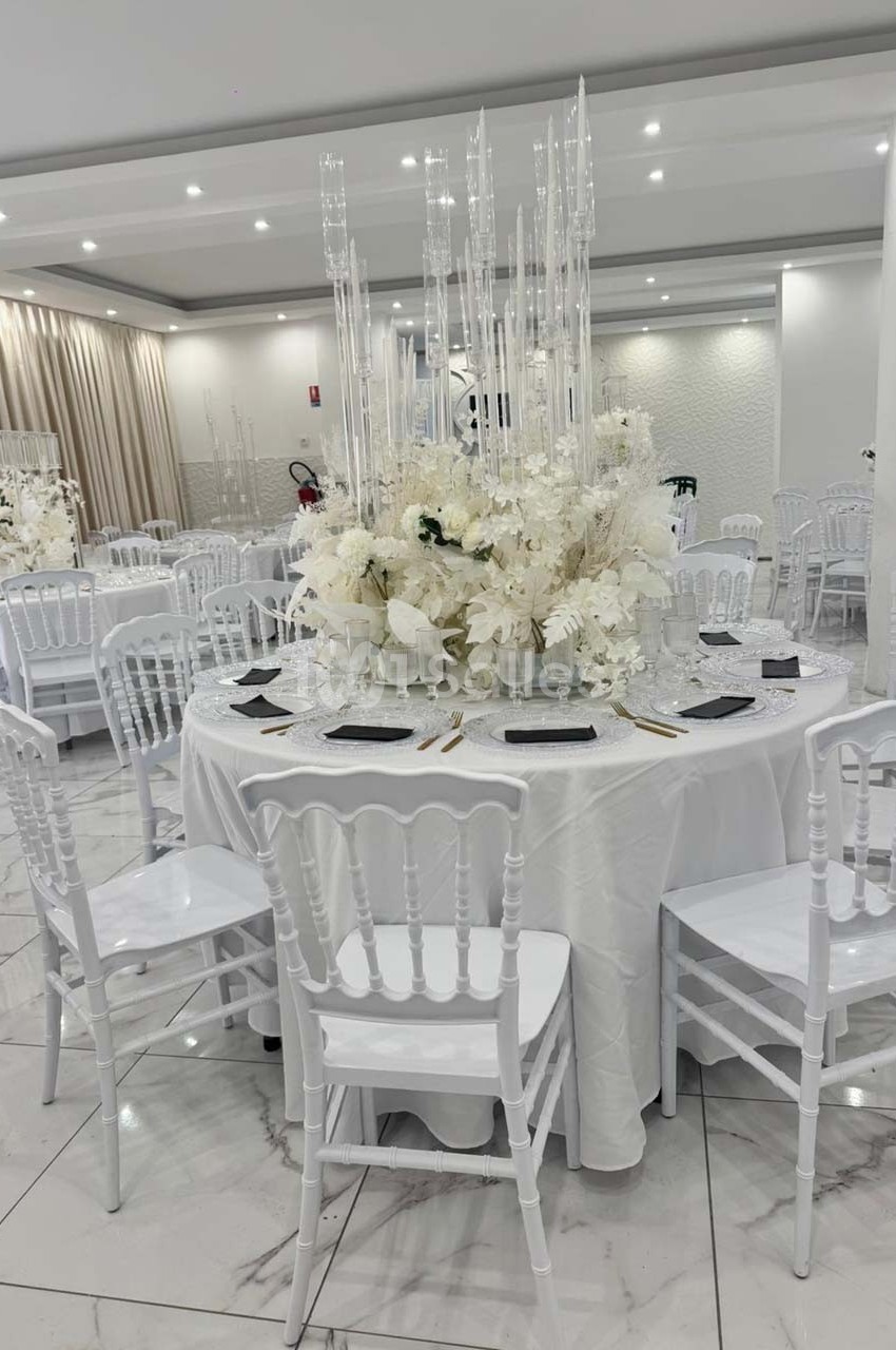 Salle de réception décorée avec des tables rondes, nappes blanches, chaises blanches et centre de table floral élégant.