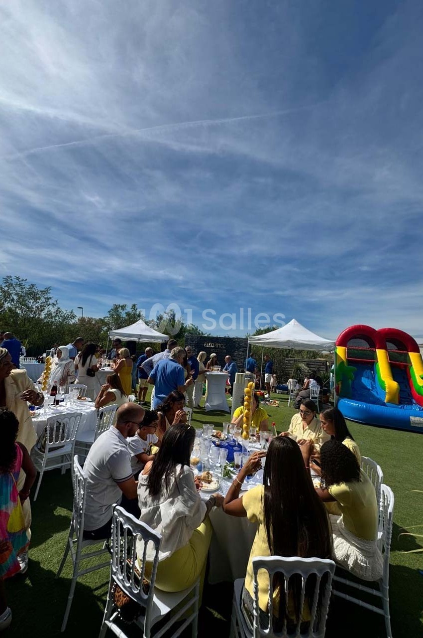 Des personnes assises et debout lors d'un événement en plein air avec des tables, des tentes blanches et un château…