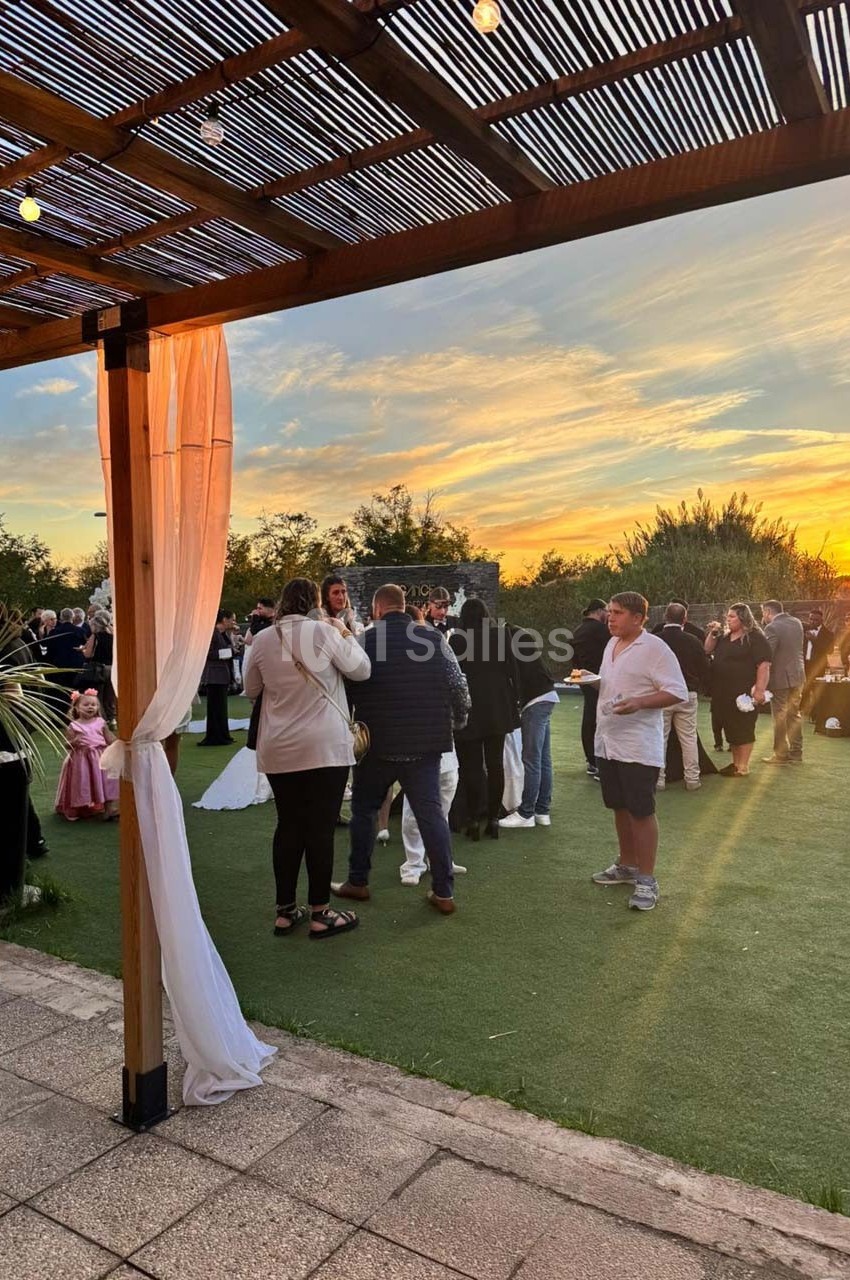 Personnes rassemblées dans un jardin au coucher du soleil, sous une pergola en bois décorée de voilages blancs.