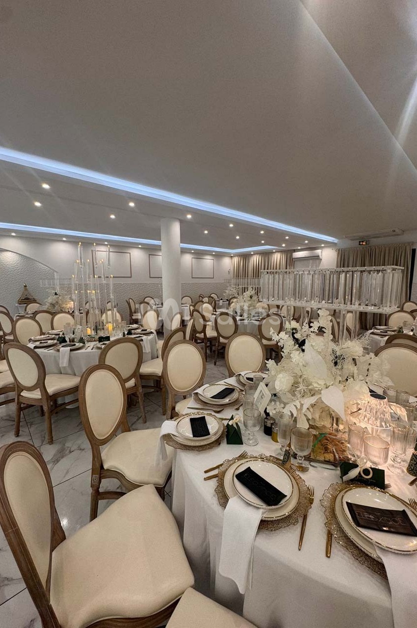 Salle de réception élégante avec tables rondes dressées, chaises en bois clair et décorations florales blanches.
