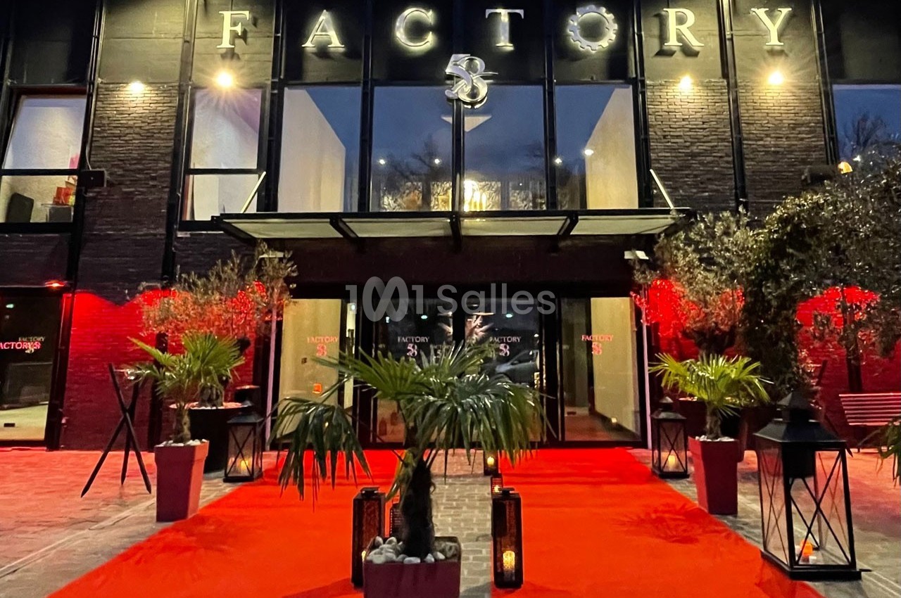 Entrée d'un bâtiment moderne avec façade noire, tapis rouge, plantes et éclairage nocturne.