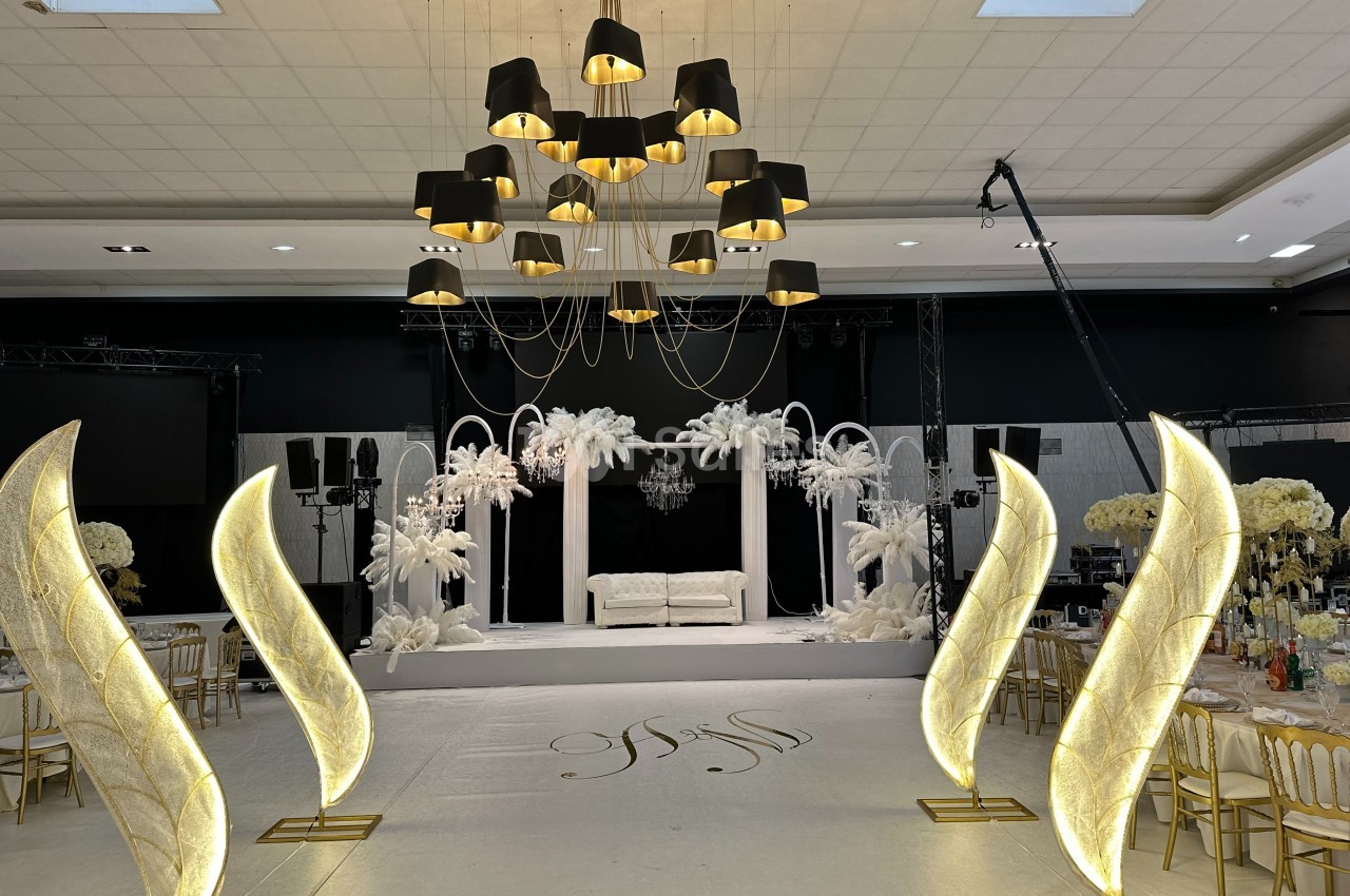 Salle de réception décorée avec des arches blanches, des palmiers artificiels et des luminaires dorés modernes.