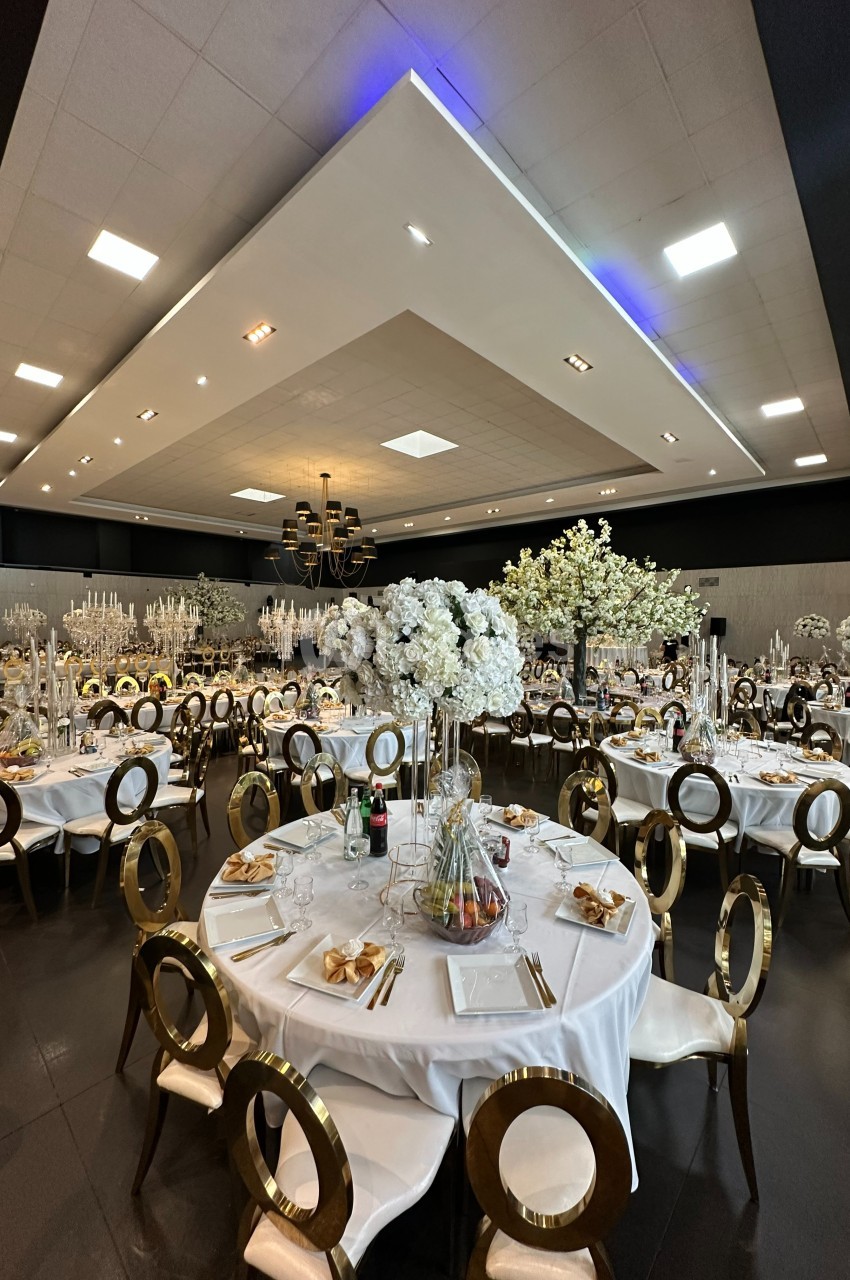 Salle de réception élégante avec tables rondes décorées de nappes blanches, chaises dorées et centres de table fleuris.