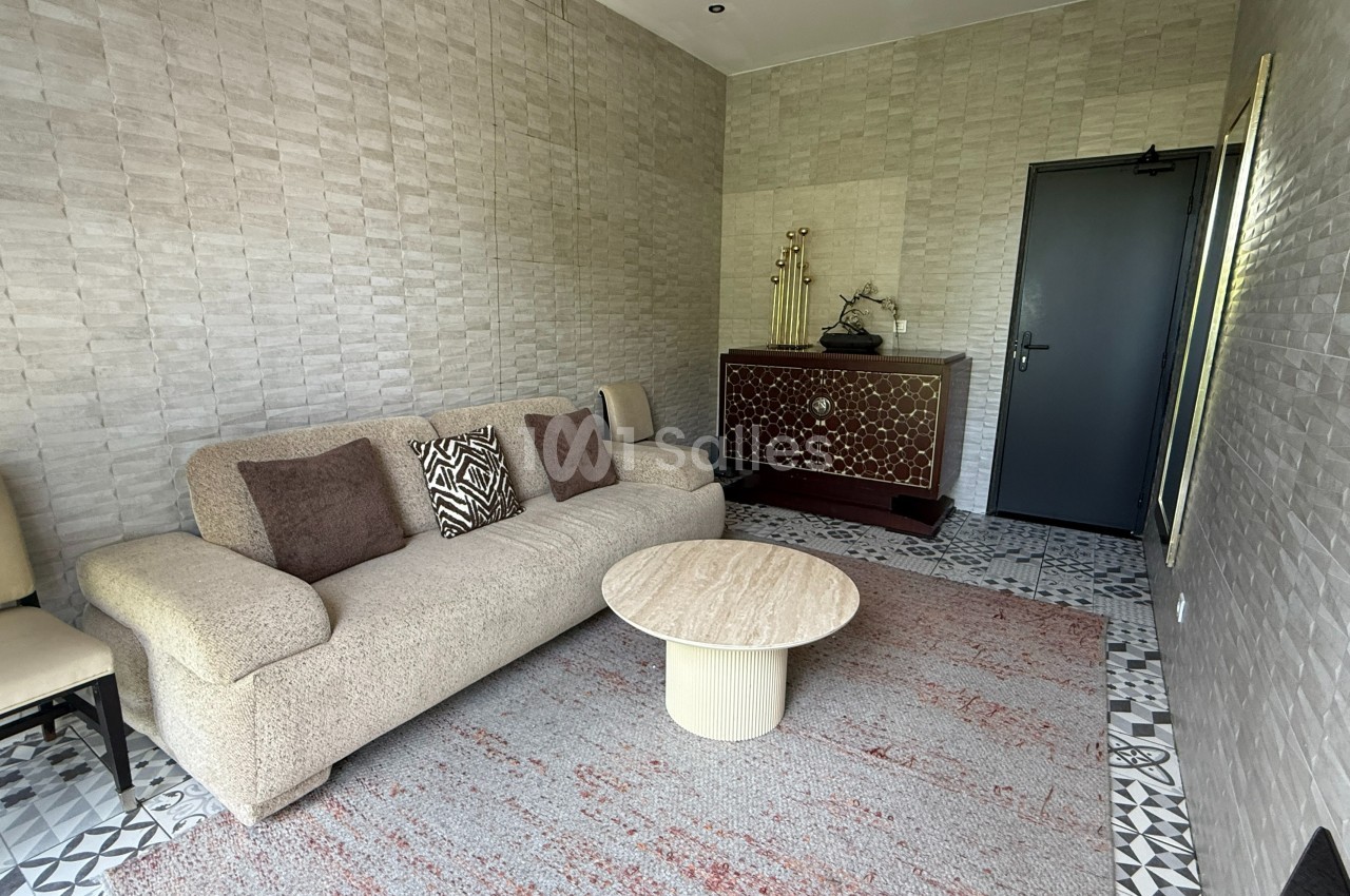 Salon avec canapé beige, table basse ronde, commode en bois sculpté et sol en carreaux décoratifs.