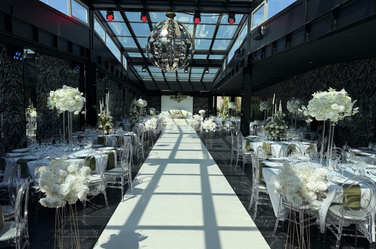 Salle de réception élégante avec tables décorées de fleurs blanches et un long tapis blanc sous une verrière.