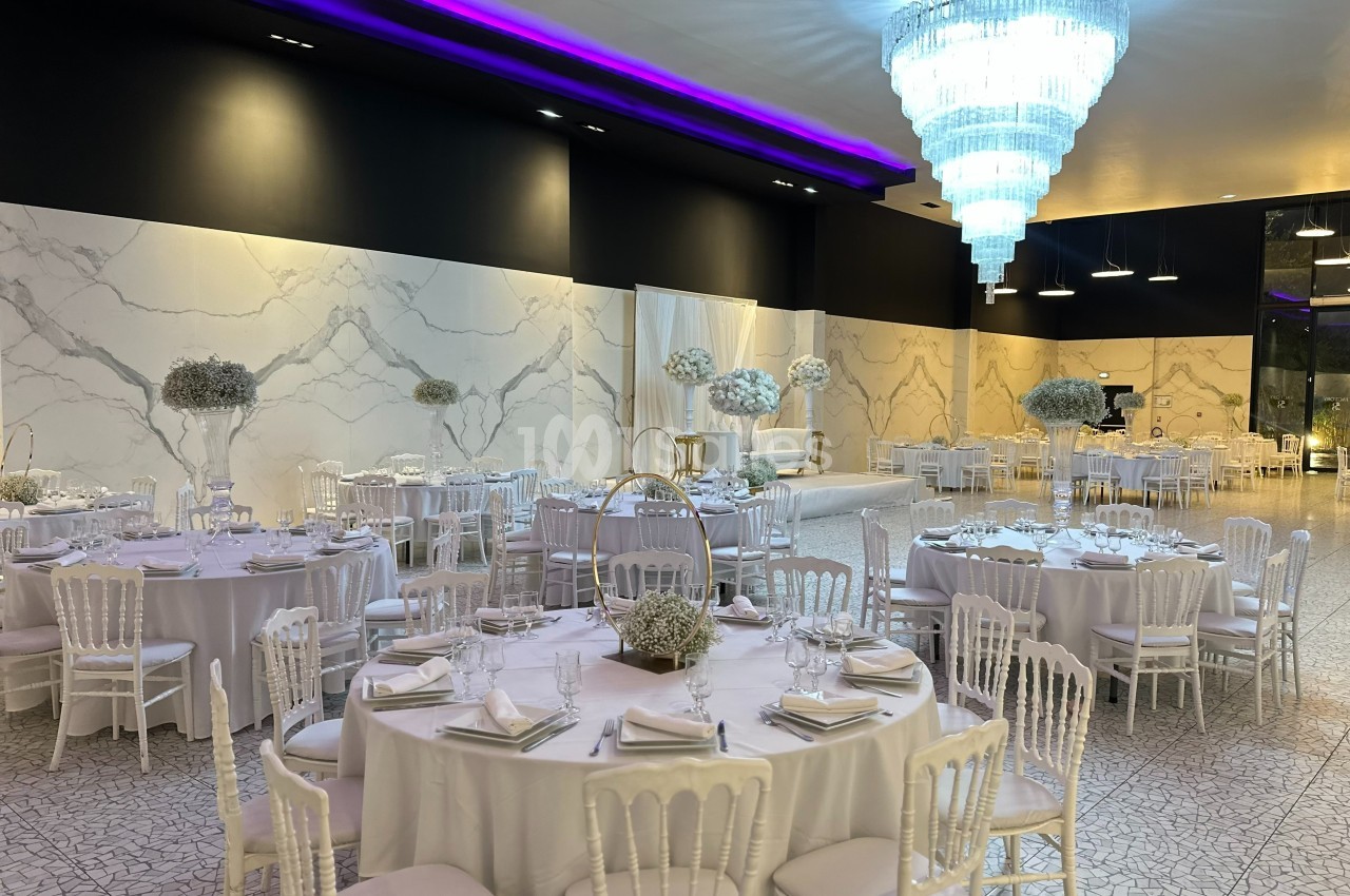 Salle de réception élégante avec tables rondes dressées, chaises blanches et grand lustre lumineux.