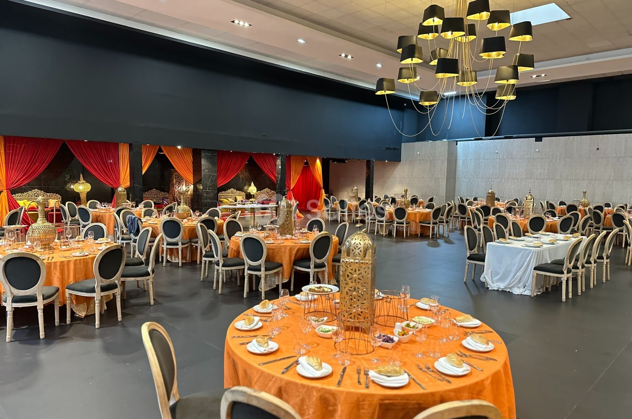 Salle de réception décorée avec des tables rondes nappées d'orange, des chaises noires et un grand lustre moderne.