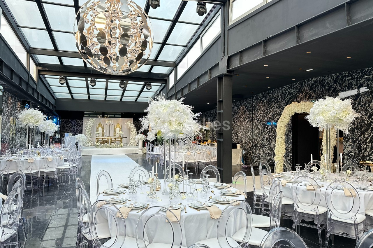 Salle de réception élégante avec tables dressées, chaises transparentes, décor floral blanc et grand lustre central.