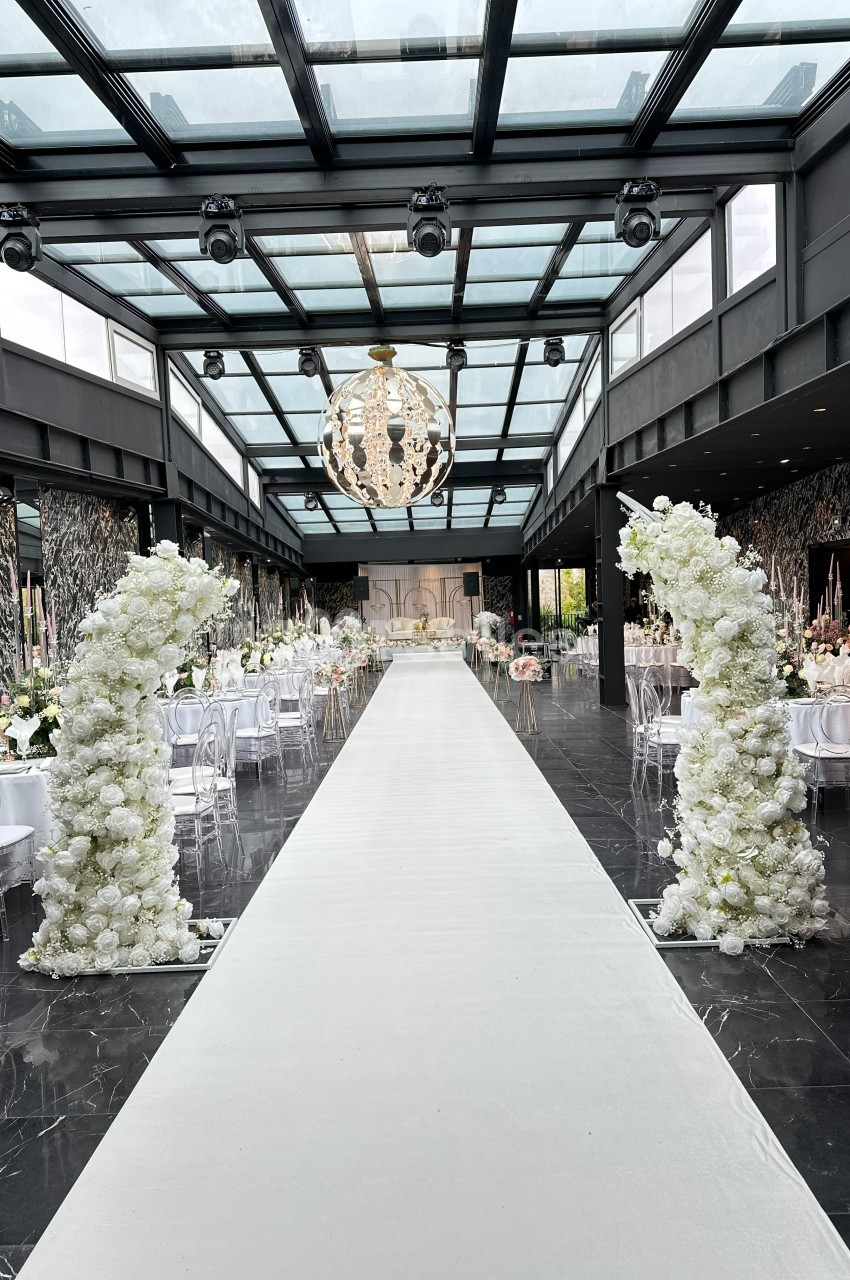 Allée centrale d'une salle de réception décorée de fleurs blanches, avec un plafond en verre et un lustre suspendu.