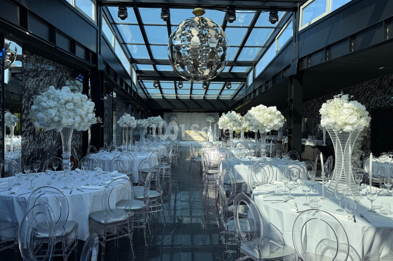Salle de réception élégante avec tables rondes dressées, chaises transparentes et décorations florales blanches.