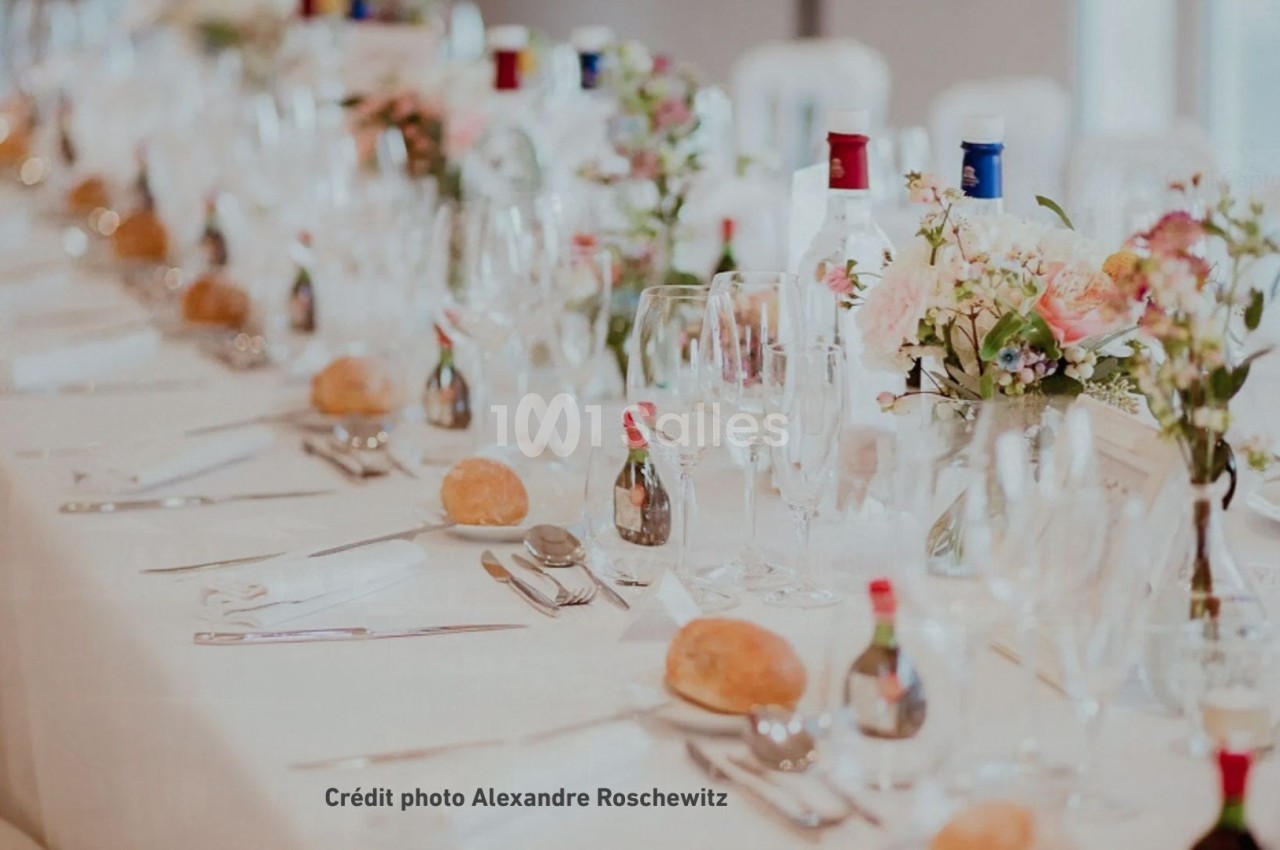 Table de réception élégamment dressée avec nappes blanches, verres, couverts, fleurs et bouteilles disposés harmonieusement.