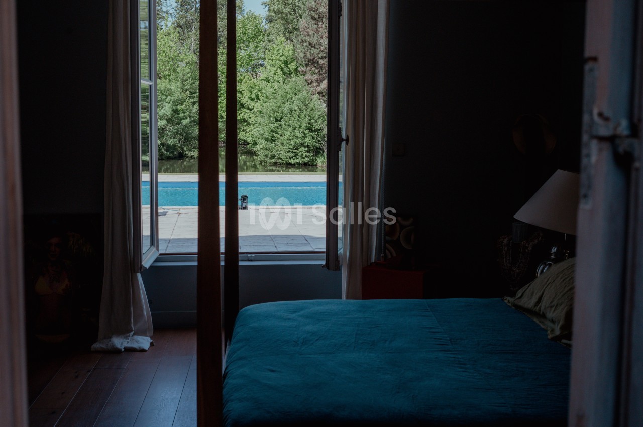 Vue depuis une chambre sombre avec lit bleu, donnant sur une terrasse et une piscine entourée de verdure.