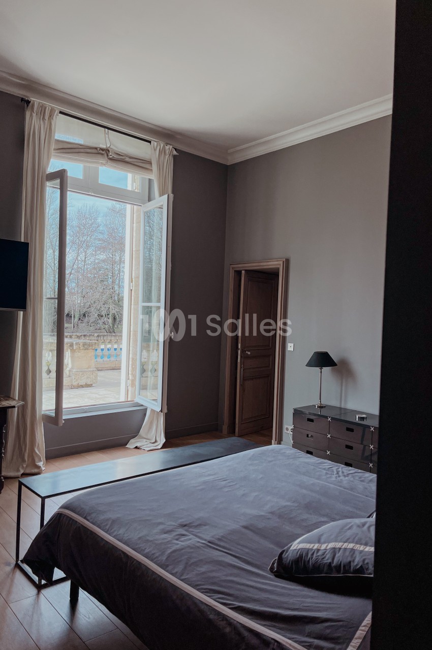 Chambre avec lit double, grande fenêtre ouverte sur un jardin, murs sombres et mobilier minimaliste.