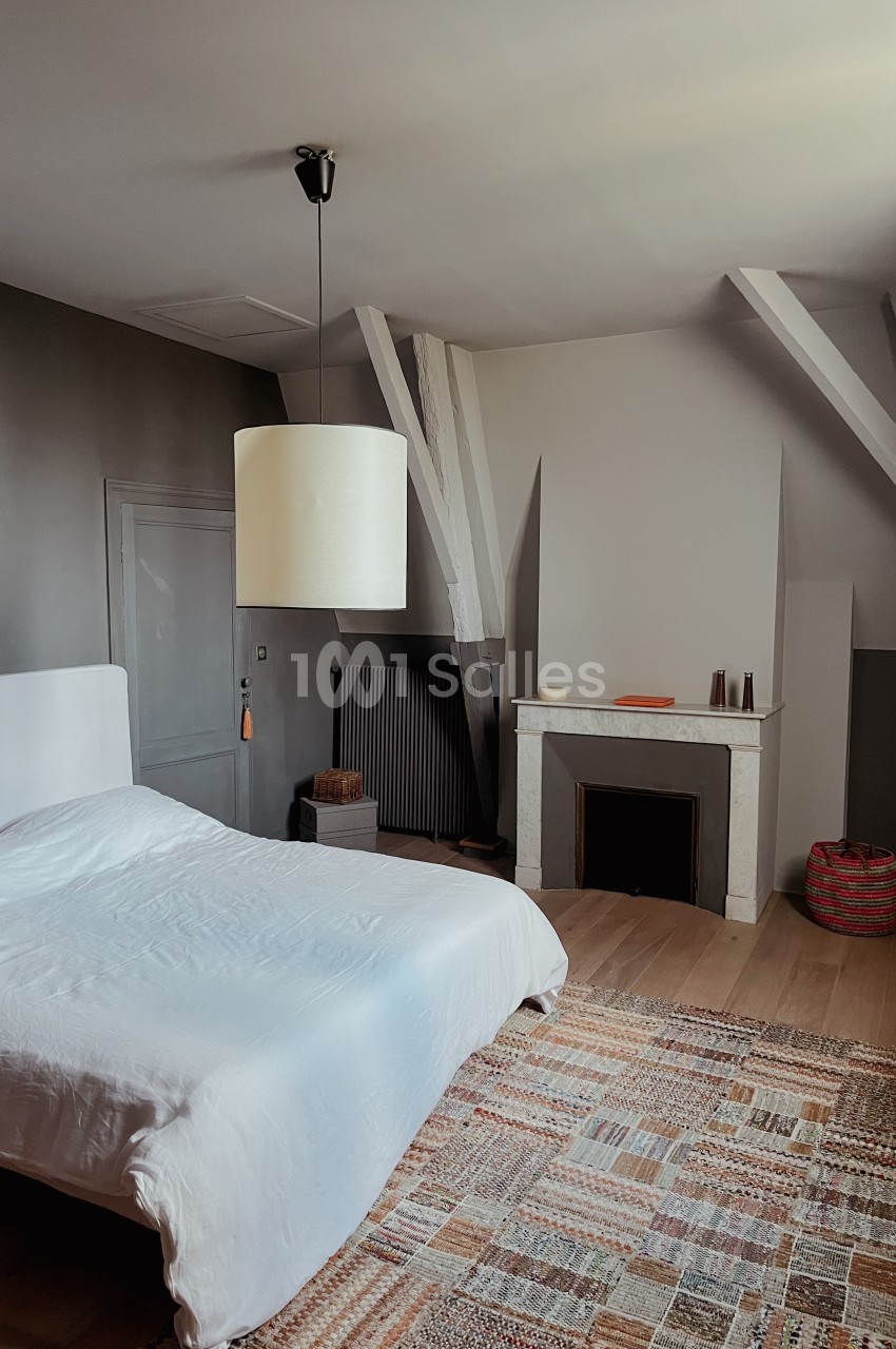 Chambre lumineuse avec lit double, cheminée décorative, tapis coloré et poutres apparentes sous un plafond mansardé.
