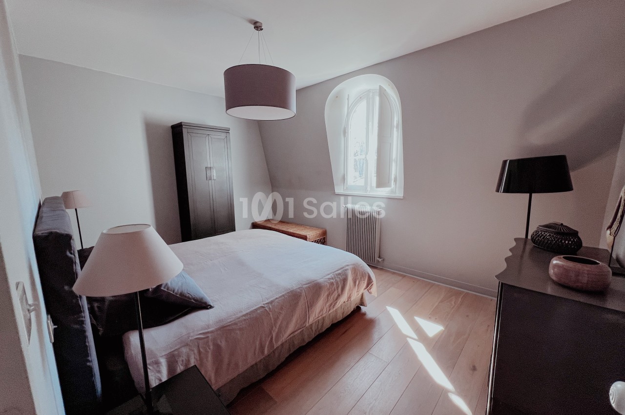 Chambre lumineuse avec lit double, armoire sombre, lampes et fenêtre cintrée laissant entrer la lumière naturelle.
