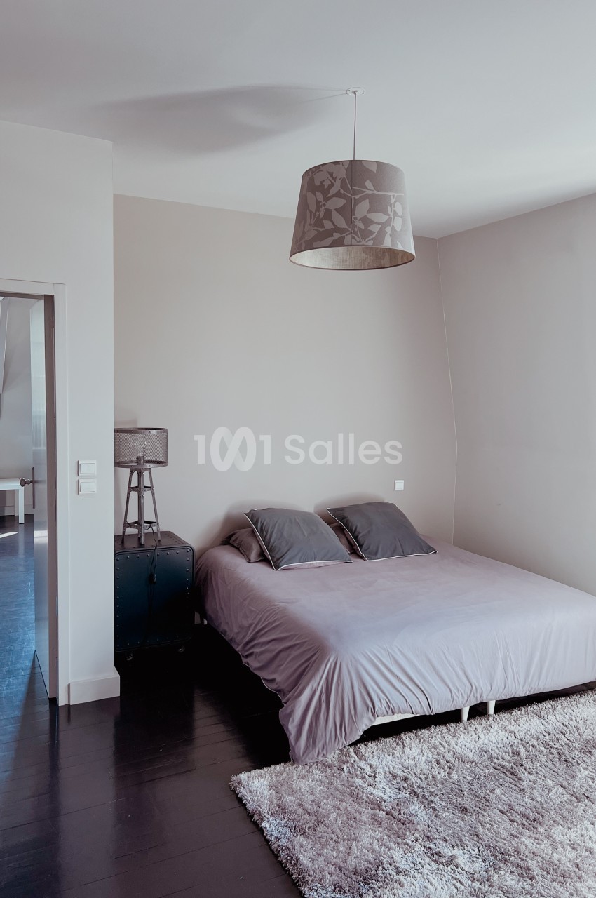 Chambre minimaliste avec lit double, murs beiges, parquet sombre, tapis clair et lampe suspendue.