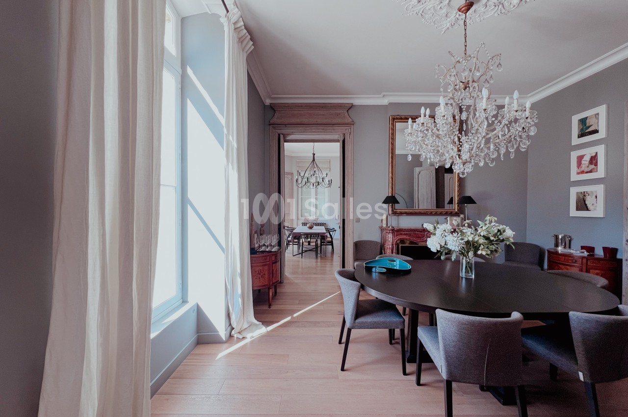 Salle à manger élégante avec table en bois, chaises grises, lustre en cristal et grande fenêtre laissant entrer la lumière.