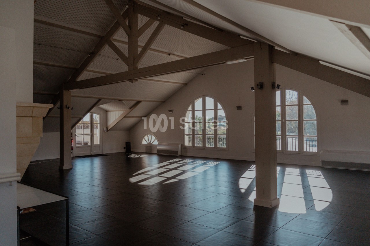 Salle spacieuse avec poutres apparentes, grandes fenêtres cintrées laissant entrer la lumière naturelle.