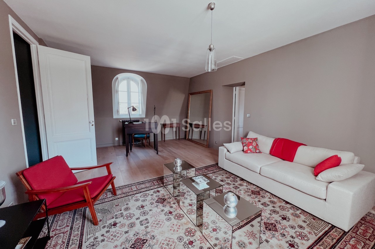 Salon lumineux avec canapé blanc, fauteuil rouge, tapis à motifs, bureau en bois et grande fenêtre cintrée.