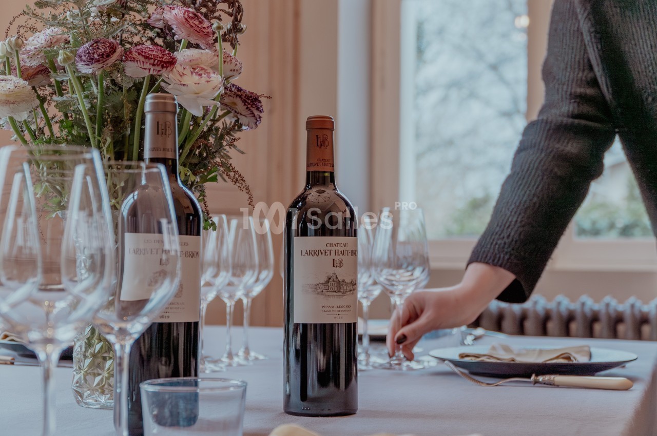 Bouteilles de vin rouge sur une table dressée avec des verres, des fleurs et une personne ajustant un couvert.