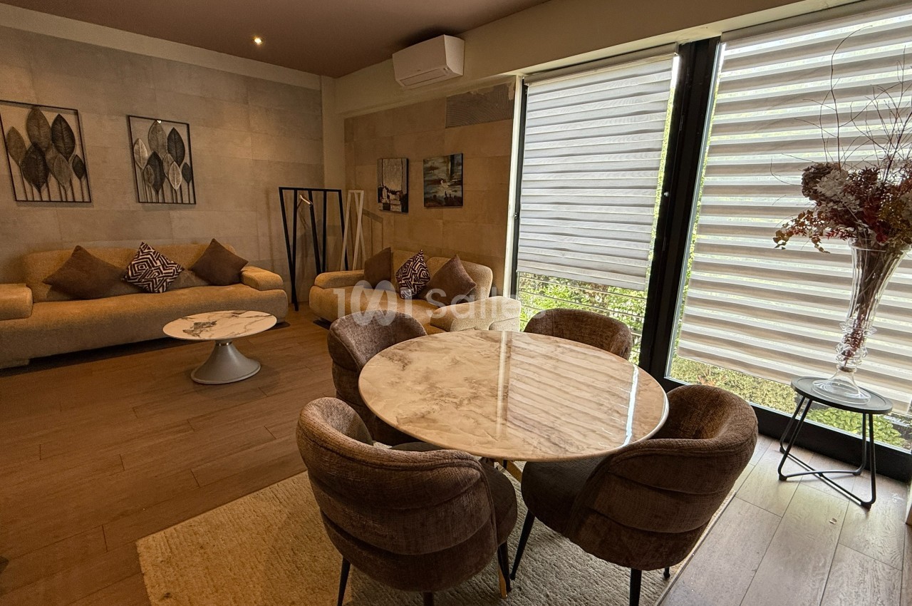 Salon moderne avec canapés beiges, table ronde en marbre, chaises rembourrées et grande fenêtre avec stores.